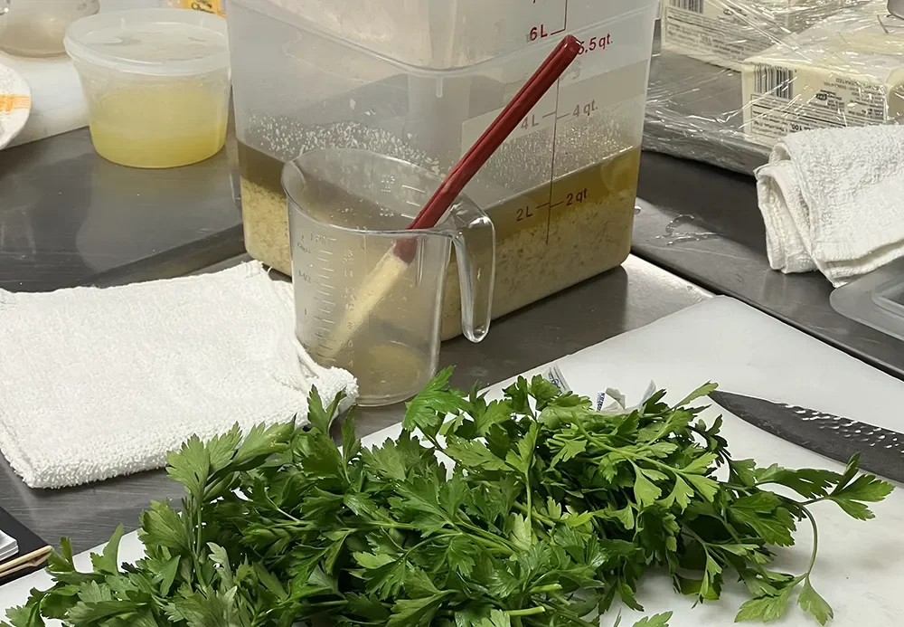 Fresh parsley on a cutting board beside containers of stock and ingredients on a stainless steel prep counter.