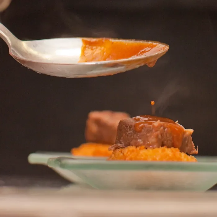 Spoon drizzling rich brown sauce over a plated piece of meat served on a bed of grains.