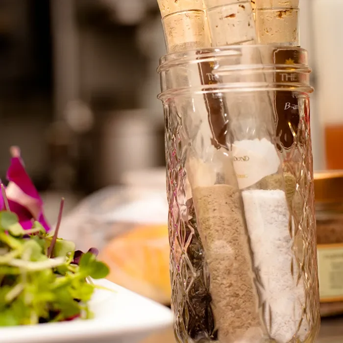 Glass jar holding assorted spices and seasonings on a kitchen counter, with a plated dish blurred in the background.