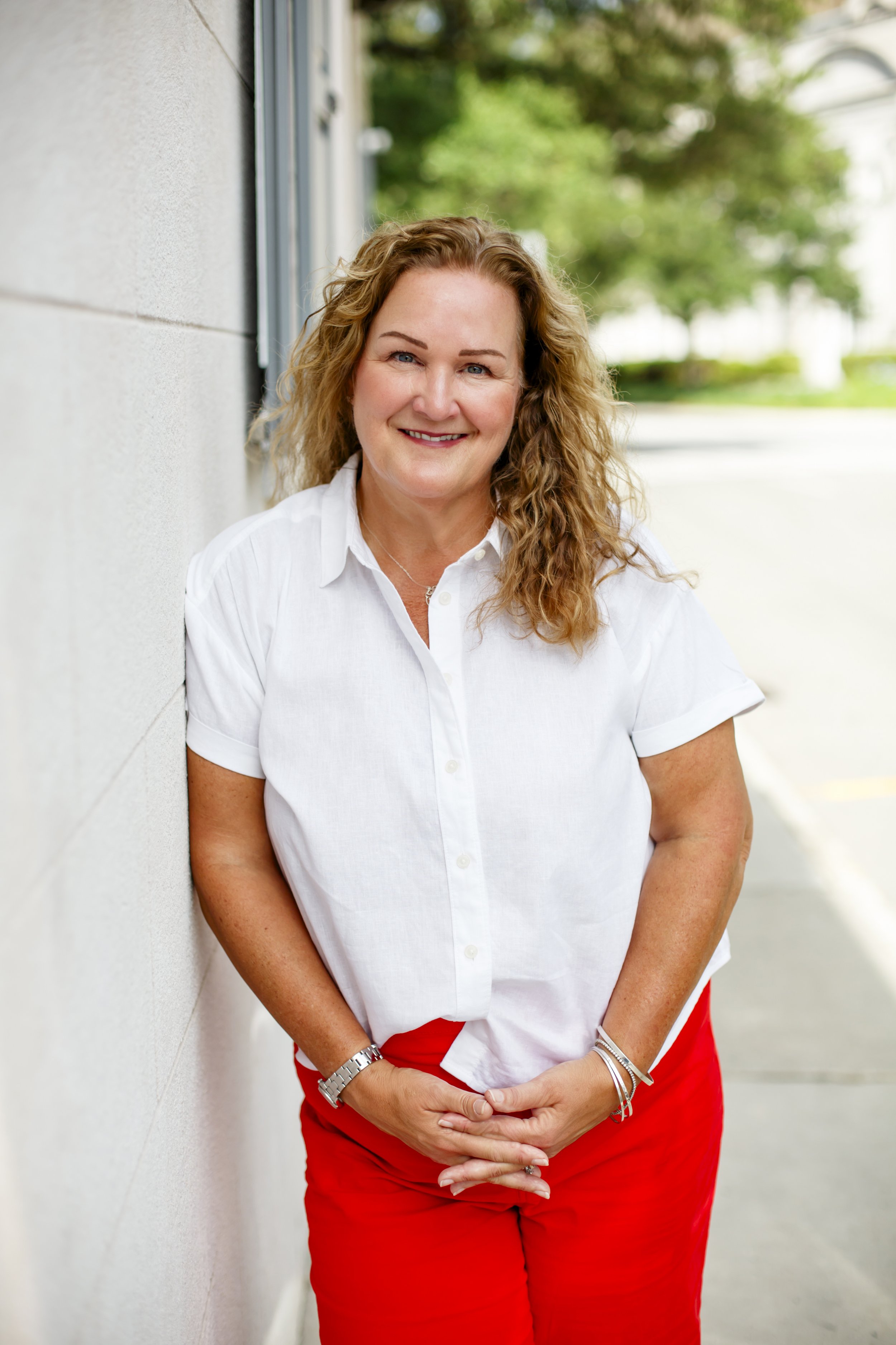 A woman with curly, blonde hair smiling, wearing a white shirt and red pants, standing outdoors leaning against a white wall with green trees blurred in the background.