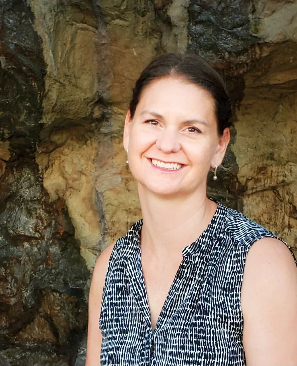 A woman smiling at a kitchen counter with a cutting board, fresh vegetables including cucumbers, a red bell pepper, and sliced carrots, and a knife.