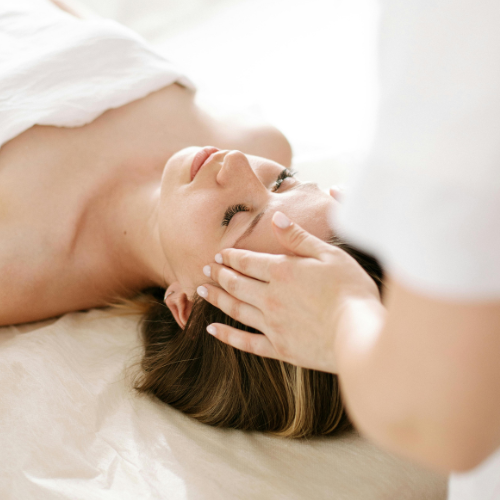 A woman receiving a facial treatment from a professional wearing gloves, lying on her back with eyes closed, in a spa or clinic setting.