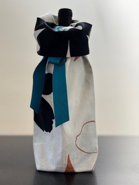 Upcycled fabric bottle bag in white with large floral pattern in black, green, tan and bright blue. Lining matches outside of bag. Bag has bright blue satin ribbon.
