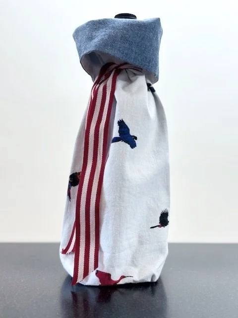 Upcycled fabric bottle bag in a pattern of flying birds on a white background. Lining is coordinating chambray blue. Bag has red and white striped ribbon.