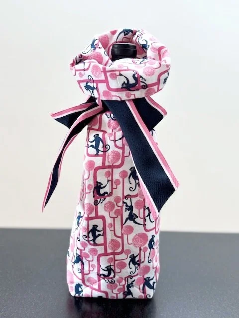 Upcycled fabric bottle bag in whimsical pattern of navy monkeys climbing on bright pink tress on a white background.Lining matches outside of bag. Bag has navy, pink and white striped ribbon.