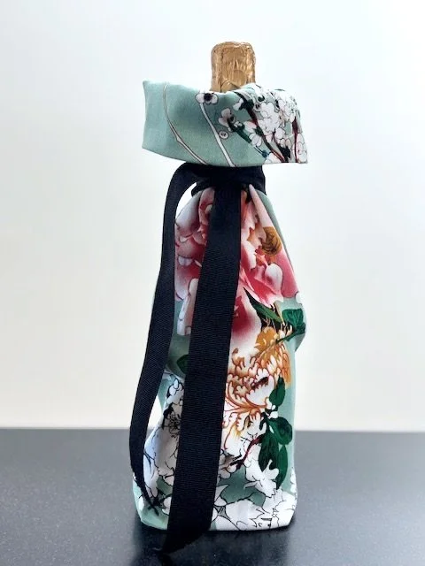 Upcycled fabric bottle bag in colorful floral pattern in shades of pink on a light teal background. Lining matches outside of bag. Bag has a black ribbon.