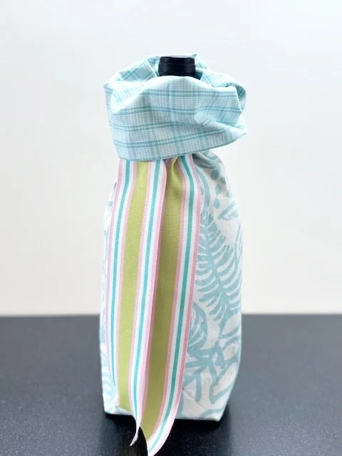Upcycled fabric bottle bag in bold tropical floral pattern in light teal blue on a white background. Coordinating lining in small teal and white check. Bag has teal, pink, green and white striped ribbon.
