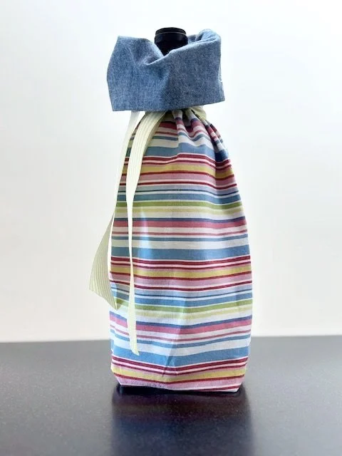 Upcycled fabric bottle bag in a horizontal multicolored stripe in shades of blue, red, pink, white, yellow. Lined in coordinating chambray blue fabric. Bag has yellow and white striped ribbon.