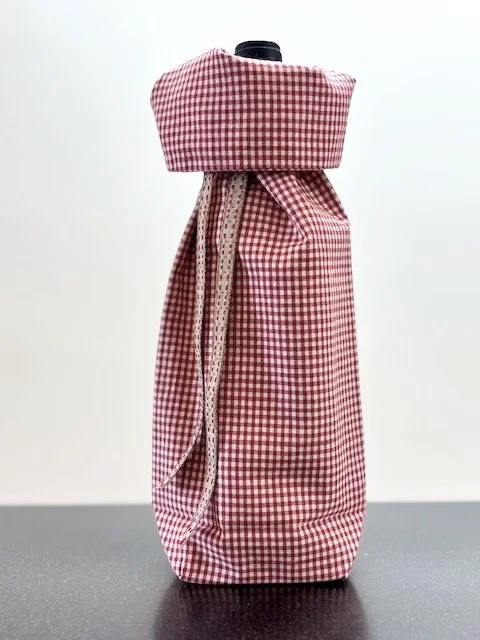 Upcycled fabric bottle bag in dark red and white gingham pattern. Lining matches outside of bag. Bag has tan and red patterned ribbon.
