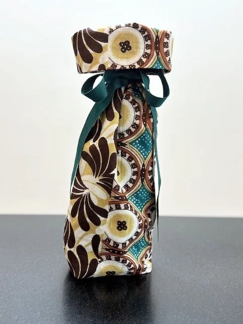 Upcycled fabric bottle bag in paneled, abstract floral pattern in shades of teal, gold, browns and white. Lining matches outside of bag. Bag has teal ribbon.