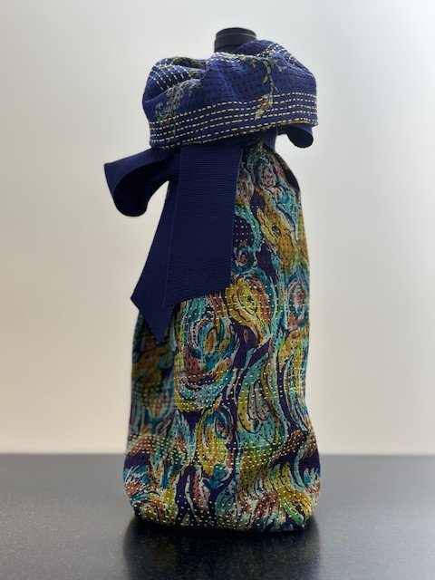 Upcycled fabric bottle bag in abstract pattern in shades of navy, teal, yellow. Bag lined in complimentary blue patterned fabric. Bag has solid navy ribbon.