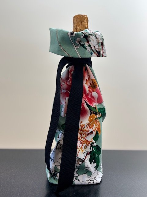 Upcycled fabric bottle bag in colorful floral pattern in shades of pink on a light teal background. Lining matches outside of bag. Bag has a black ribbon.