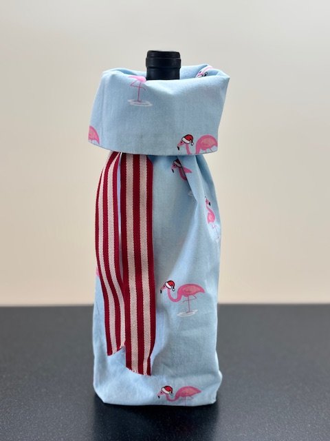 Upcycled fabric bottle bag in whimsical pattern of pink flamingos with Santa hats on a light blue background. Lining matches outside of bag. Bag has red and white striped ribbon.