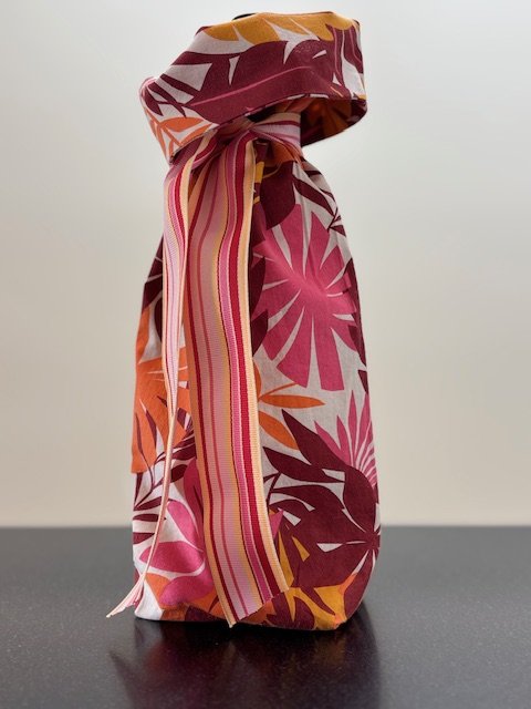 Upcycled fabric bottle bag in bold tropical floral pattern in shades of pink, orange, burgundy on a white background. Lining matches outside of bag. Bag has striped ribbon in the same colors as the bag.