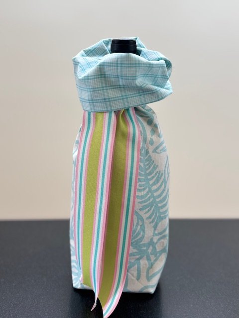 Upcycled fabric bottle bag in bold tropical floral pattern in light teal blue on a white background. Coordinating lining in small teal and white check. Bag has teal, pink, green and white striped ribbon.