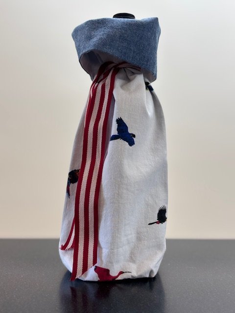 Upcycled fabric bottle bag in a pattern of flying birds on a white background. Lining is coordinating chambray blue. Bag has red and white striped ribbon.