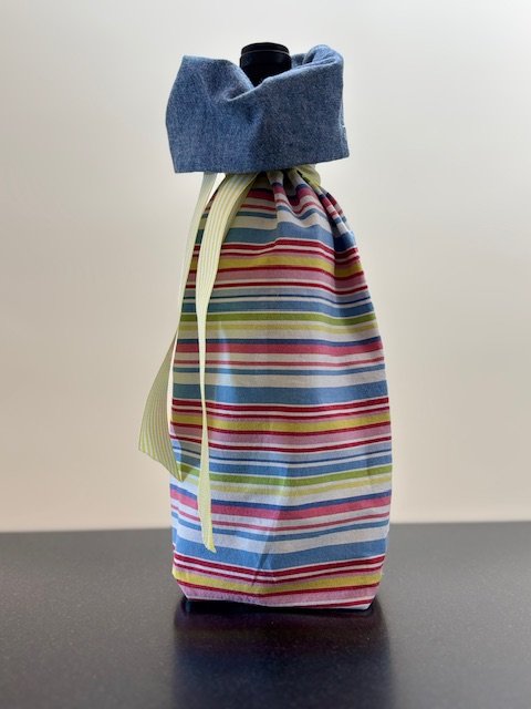 Upcycled fabric bottle bag in a horizontal multicolored stripe in shades of blue, red, pink, white, yellow. Lined in coordinating chambray blue fabric. Bag has yellow and white striped ribbon.