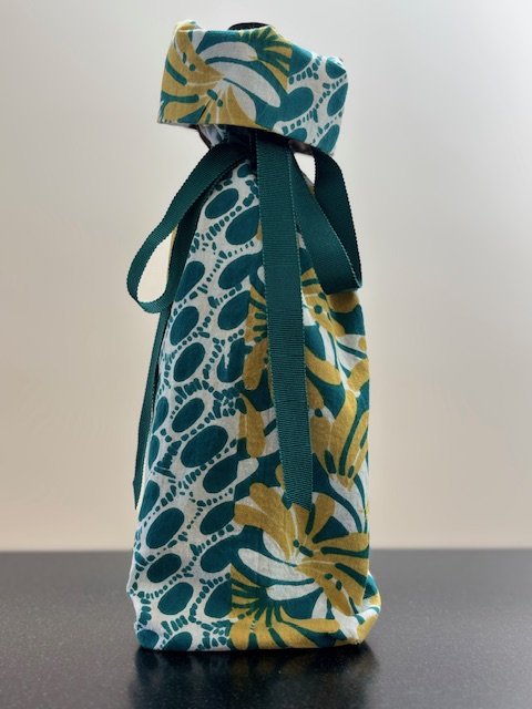 Upcycled fabric bottle bag in variety of abstract floral patterns in shades of teal, golden yellow, whites and browns . Lining matches outside of bag. Bag has teal green ribbon.