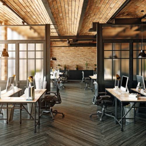 Modern office space with multiple desks, computers, and office chairs, separated by glass partitions, with a brick wall and ceiling, wooden flooring, and warm lighting.