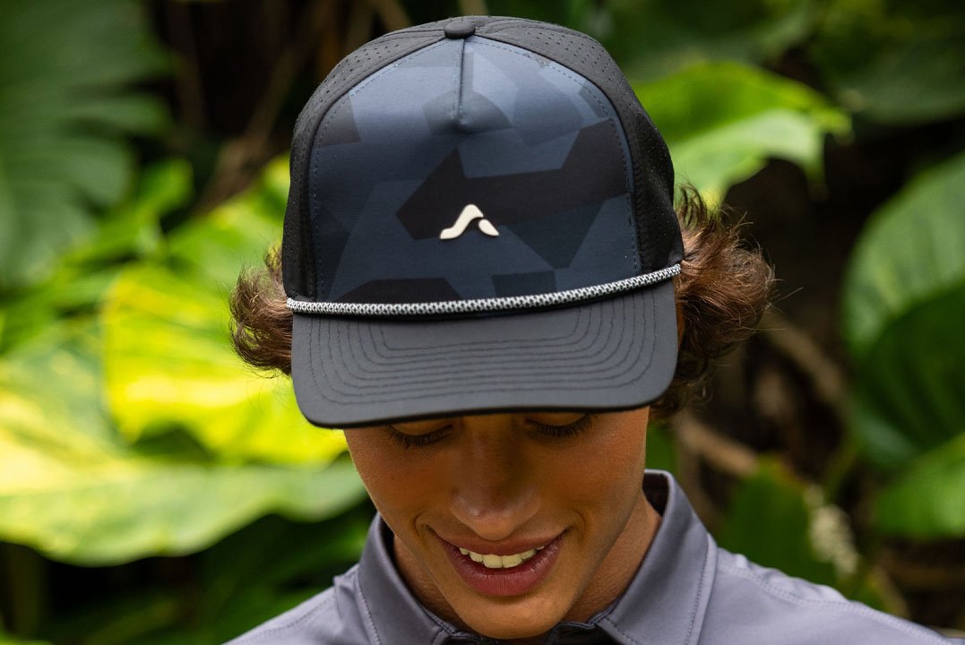 Close-up of a person wearing a black and gray baseball cap with a logo on the front, outdoors surrounded by green foliage.