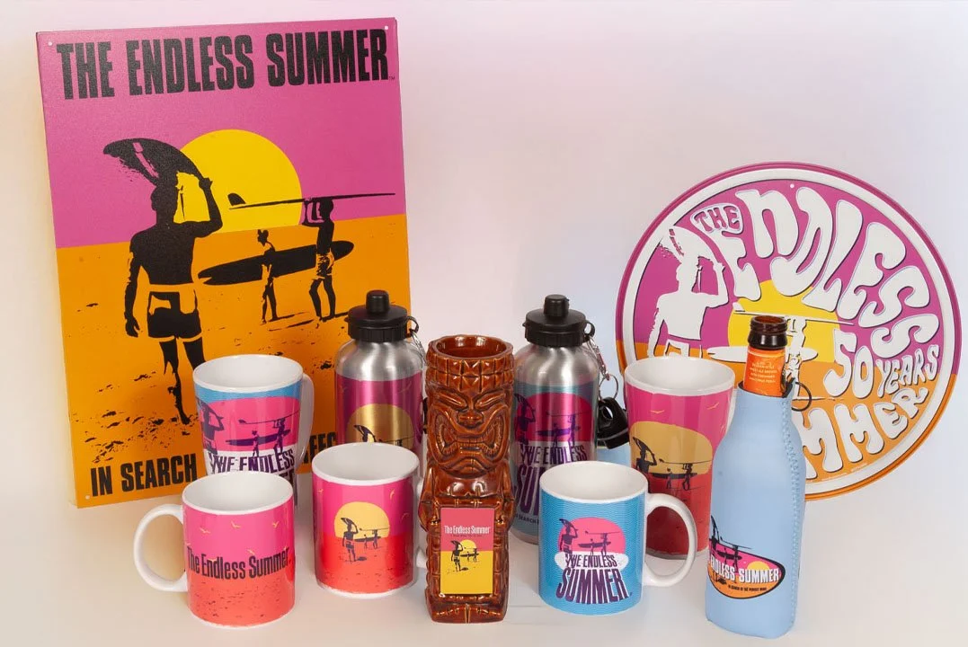 Collection of merchandise and drinkware with 'The Endless Summer' and 'In Search of the Perfect Wave' themes, including mugs, water bottles, and a tiki figurine, with posters in the background.