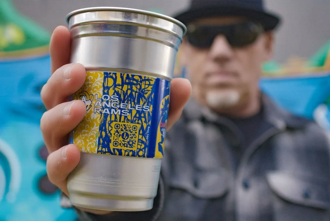 A person holding a Los Angeles Rams-branded tumbler cup with a QR code, with a person wearing sunglasses and a jacket in the background.