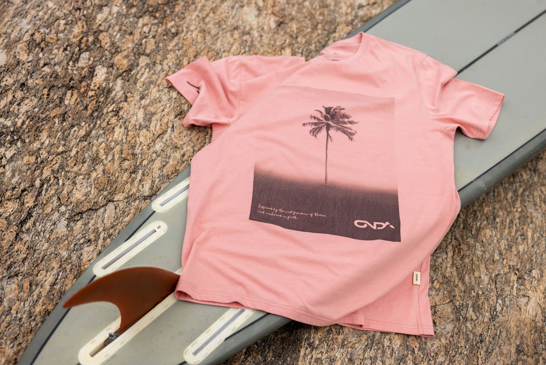 A pink T-shirt with a palm tree graphic and text, placed on a surfboard on a granite surface.