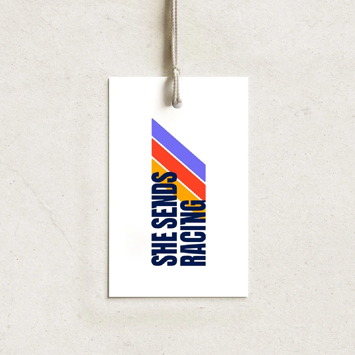 A vertical white tag with a multicolored striped design at the top, featuring blue, red, and yellow colors. The text "The Seuds Racing" is printed vertically in bold blue letters over the stripes, hanging from a white string against a neutral background.