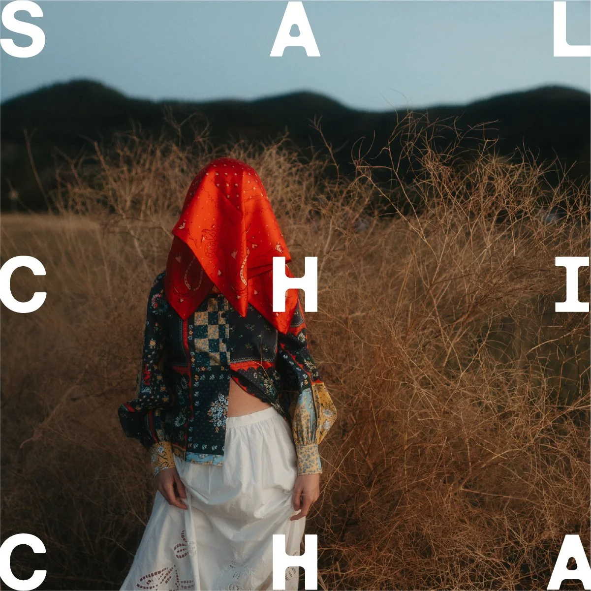 Image of a person standing in a dry field with hills in the background. The person is wearing a patterned shirt, a white skirt, and has a red bandana over their face, obscuring their identity. Large white letters spell out the word 'SACHIA' across the image.