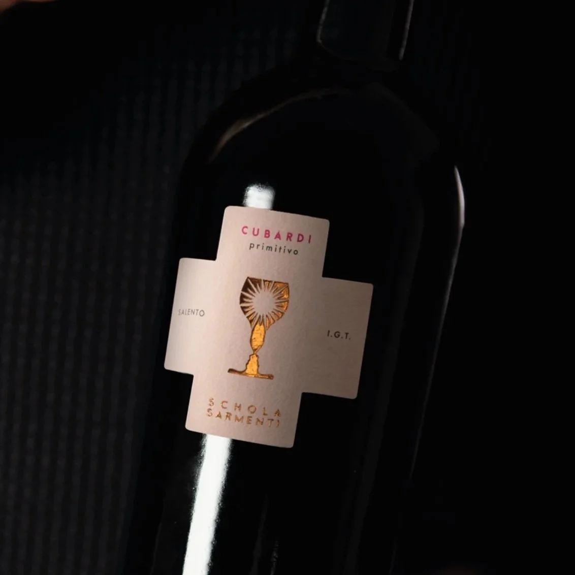A dark wine bottle with a white label featuring a gold chalice illustration, pink and gold text, and a black background.