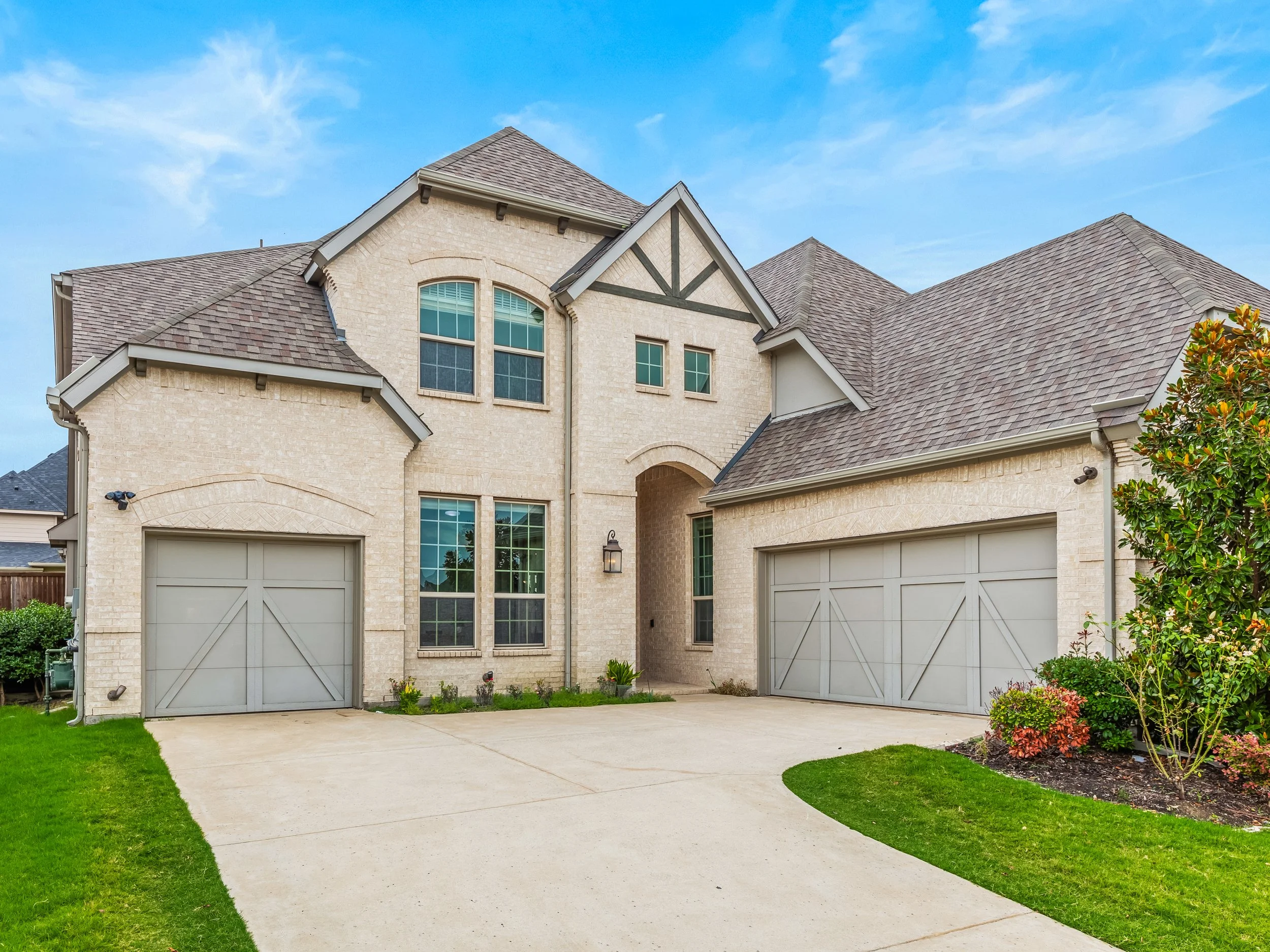 Front view of a modern two-story house with beige brick exterior, gray garage doors, and a gabled roof, with green lawn and landscaping.