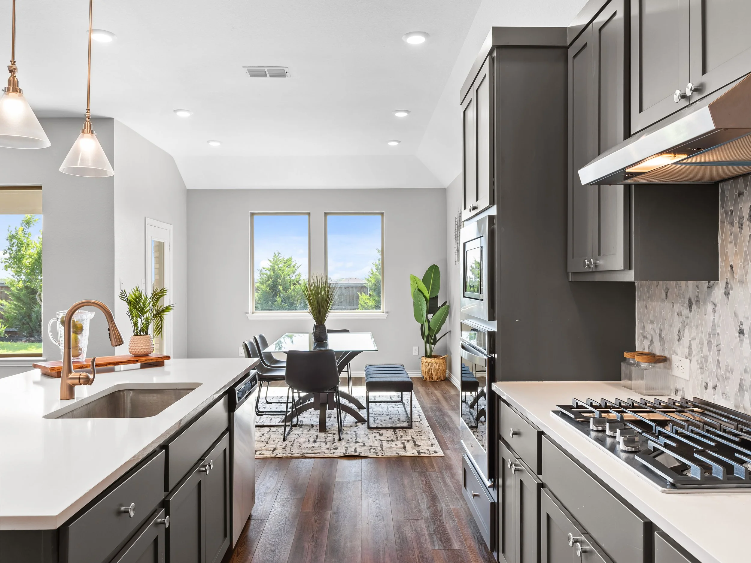 Modern kitchen with gray cabinets, white countertops, a gas stove, and a dining area with a table, chairs, and large windows showing greenery outside.