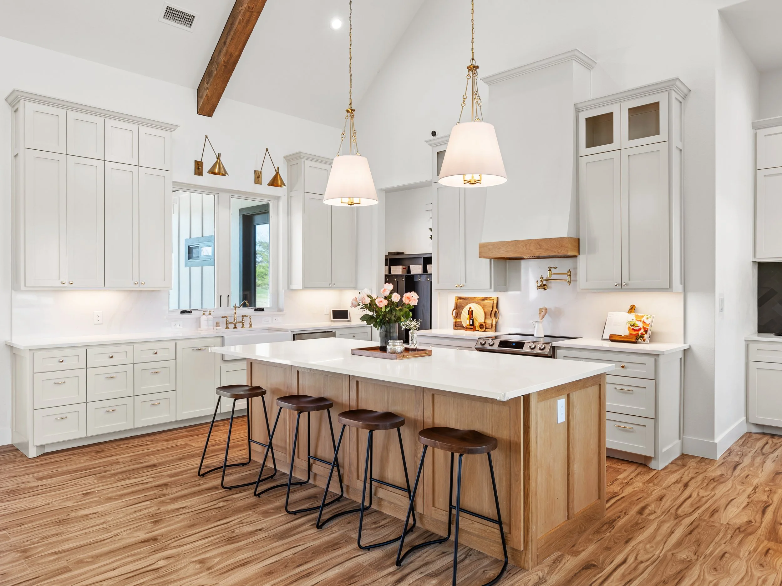 Modern kitchen with white cabinets, a wooden island with four bar stools, and hardwood floors, decorated with pendant lights and flowers on the island.