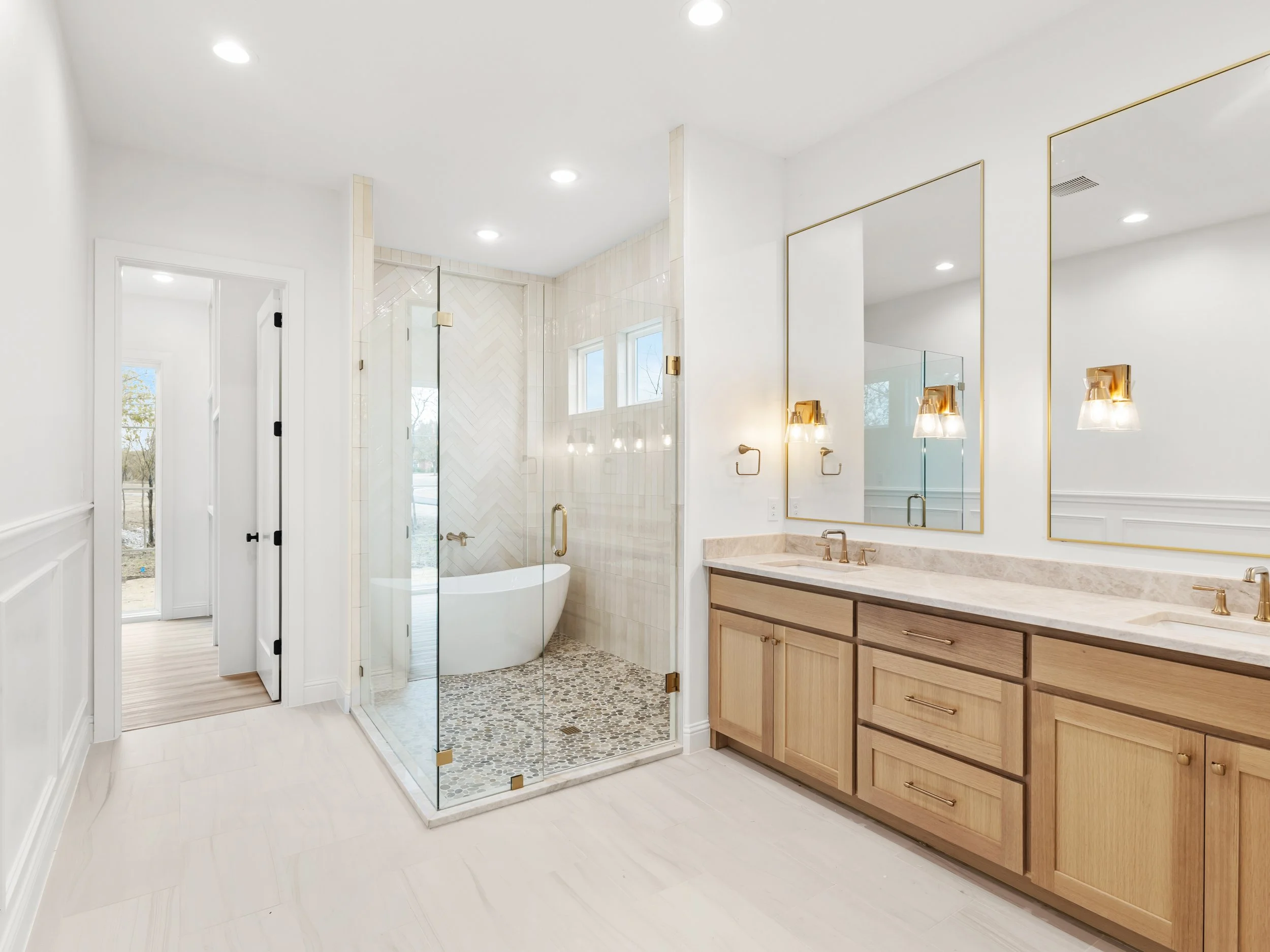 Modern bathroom with double vanity, large mirrors, beige and gold accents, glass-enclosed shower, and freestanding bathtub, with natural light coming through windows.
