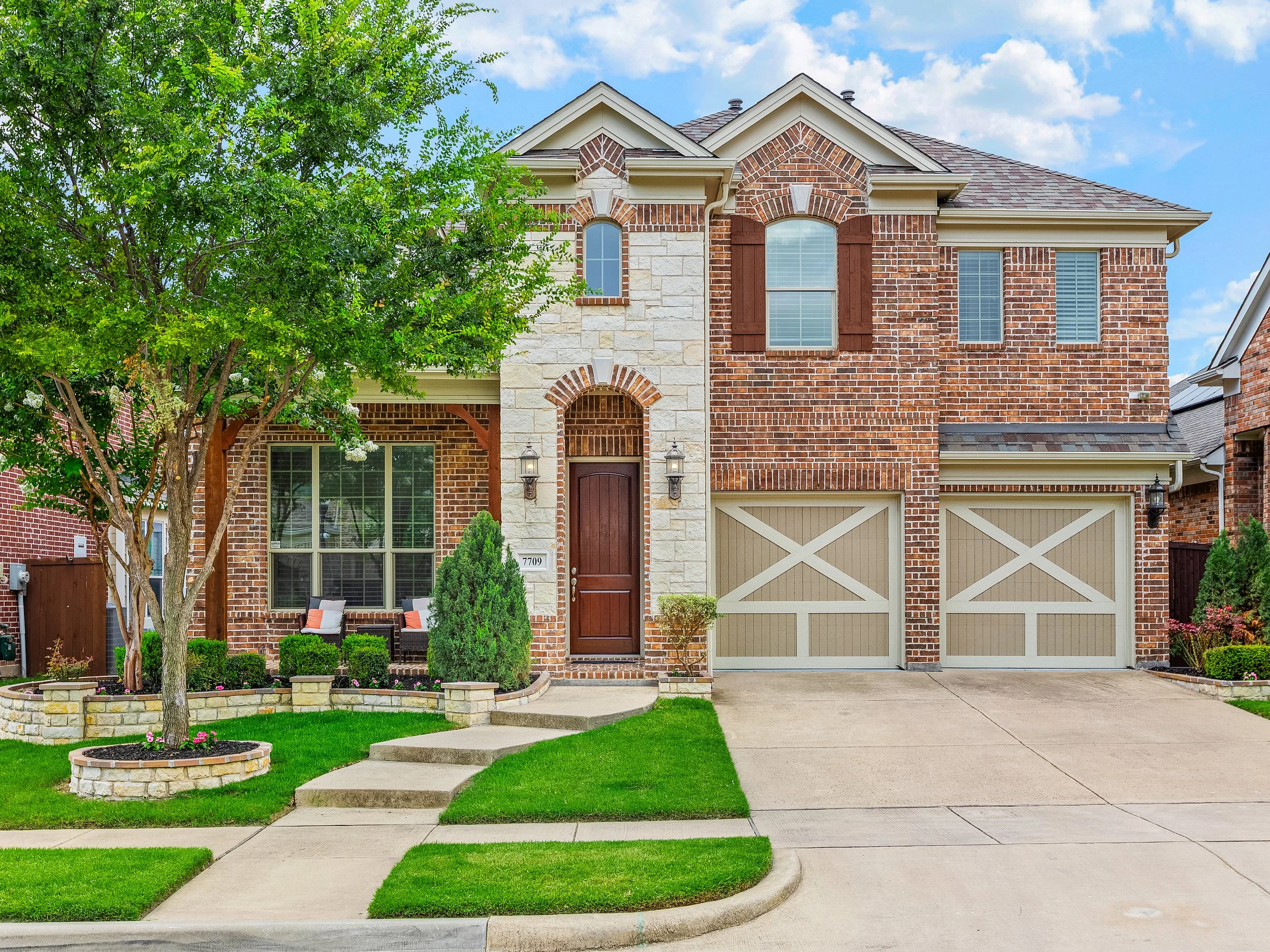 Two-story brick house with a front yard, green lawn, and sidewalk, featuring a two-car garage, wooden front door, and surrounded by trees and plants.
