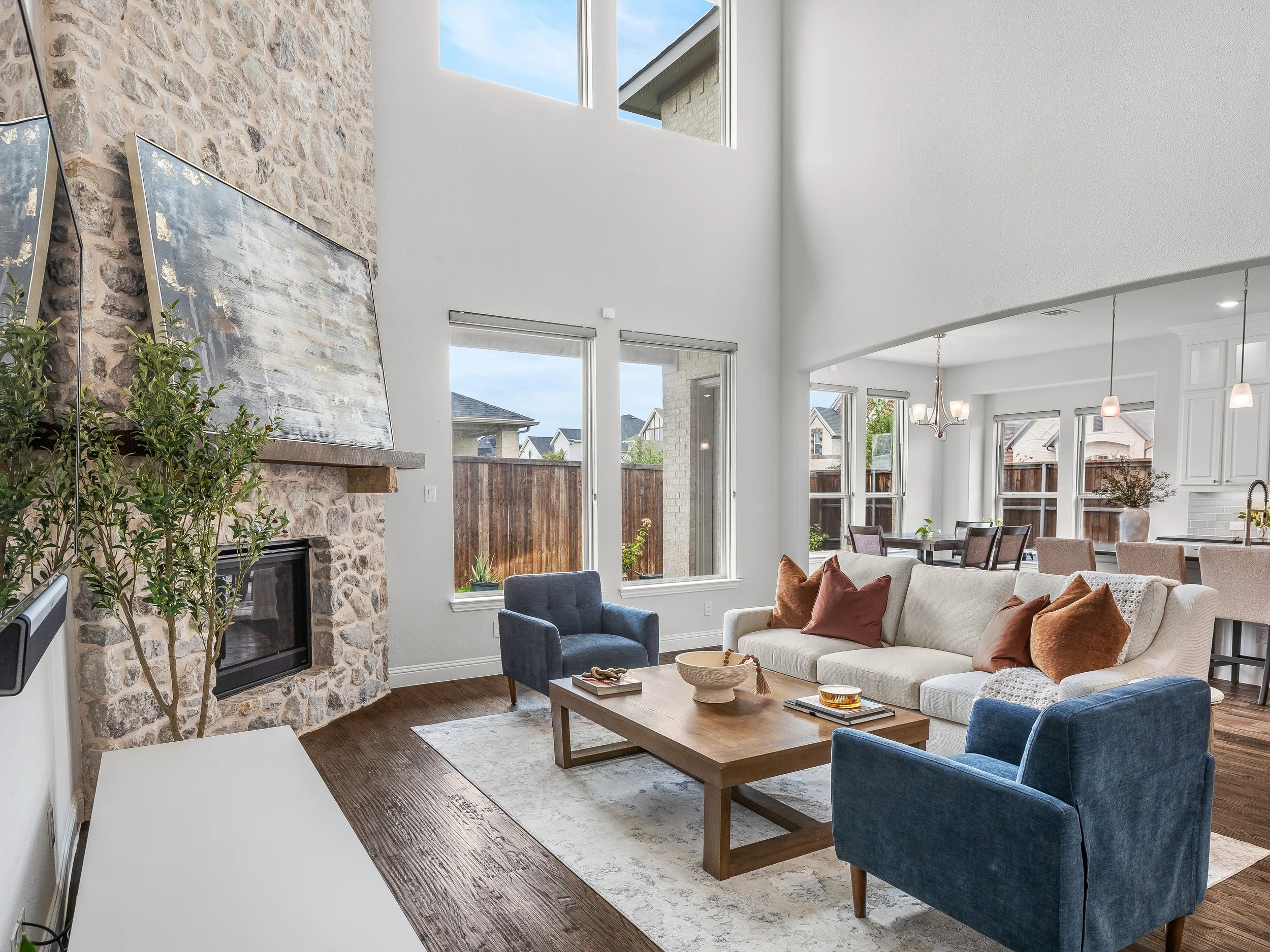 Living room with a stone fireplace, white sofa with brown and white pillows, blue armchairs, wooden coffee table, large windows, and an open dining area in the background.