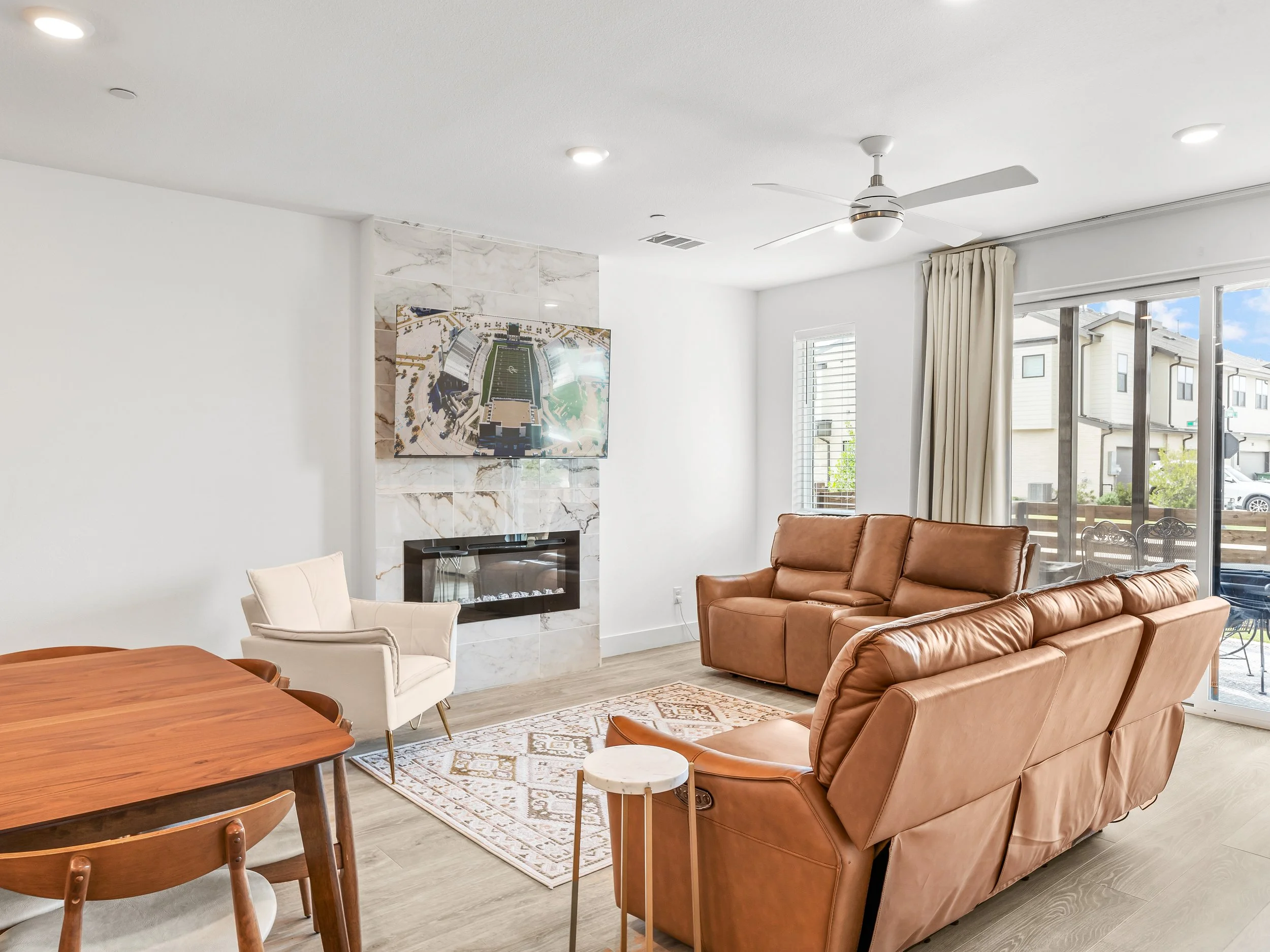 Living room with brown leather sofas, a white armchair, a wood dining table with chairs, a marble fireplace with a TV above, a window, curtains, and a sliding glass door leading outside with patio furniture.