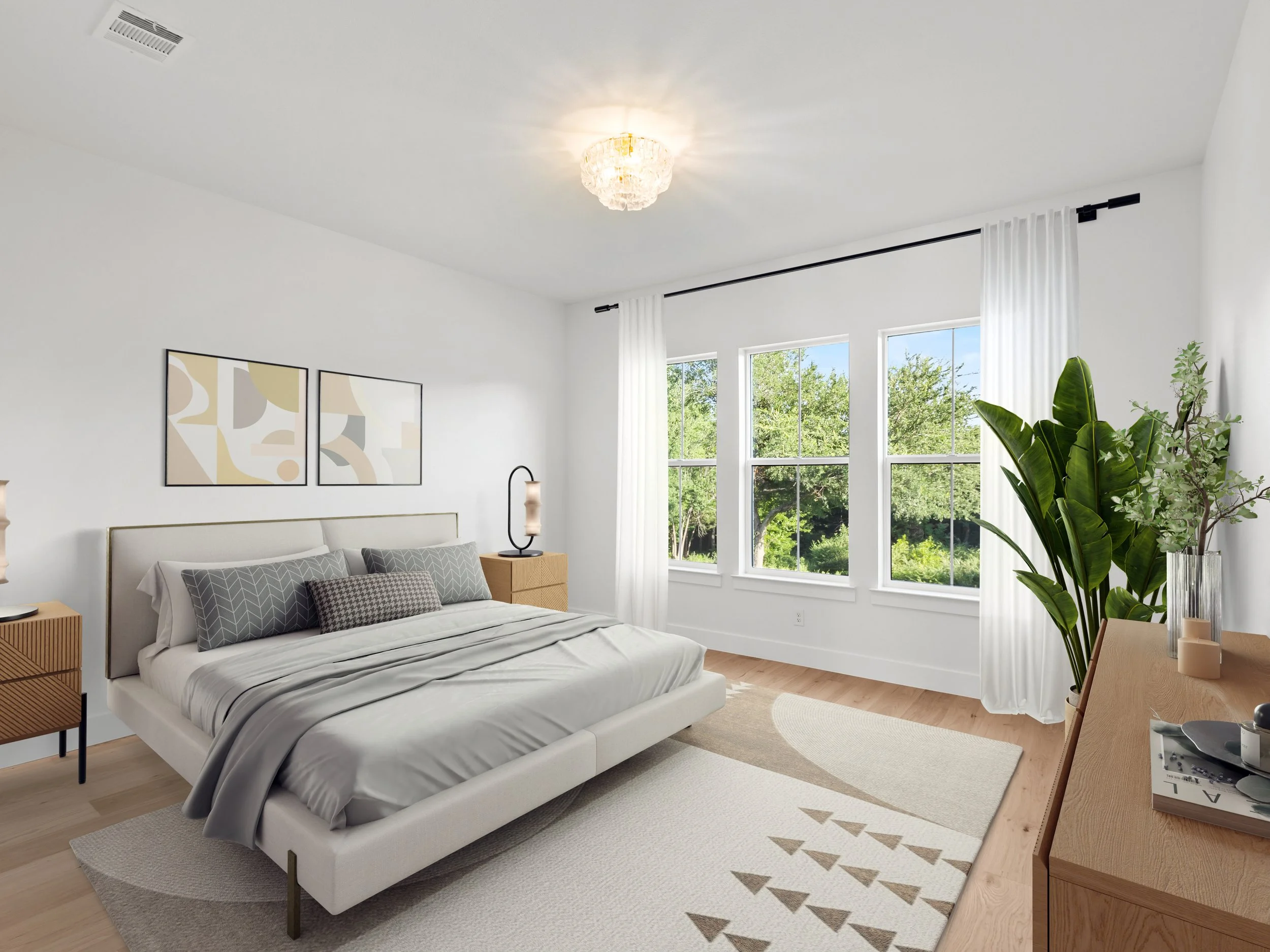 A bedroom with a white bed, gray pillows, wooden nightstands, white curtains, large windows showing greenery outside, a large green plant, and framed abstract art on the wall.