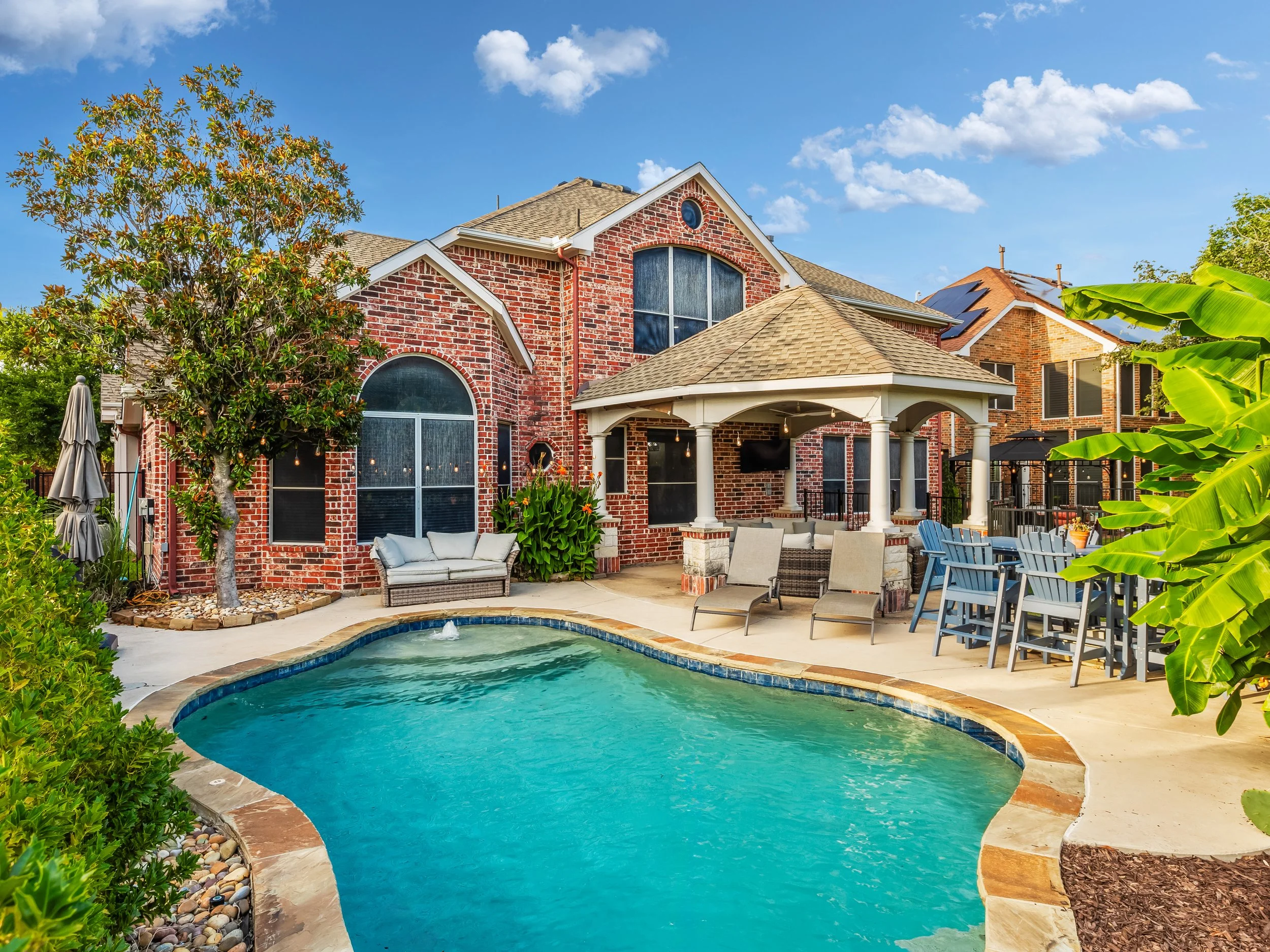 Backyard with a swimming pool, outdoor furniture, a gazebo, and a brick house with large windows under a blue sky with scattered clouds.