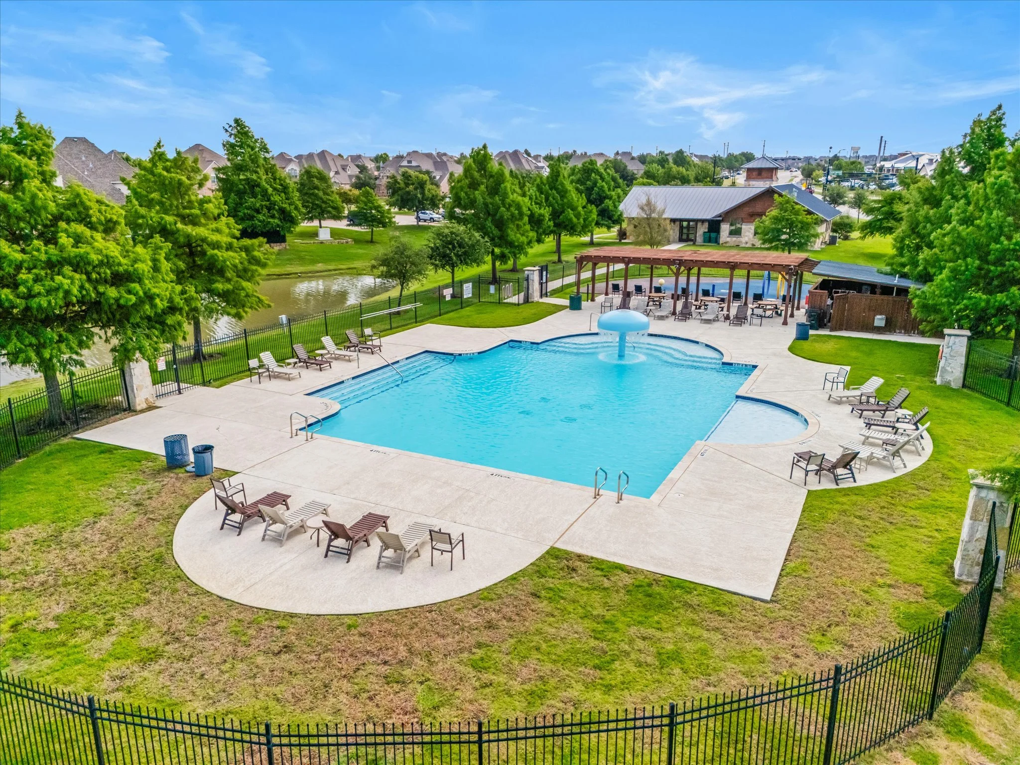 An aerial view of a community swimming pool with surrounding chairs, a covered seating area, a small pond, green trees, and a parking lot in the background.