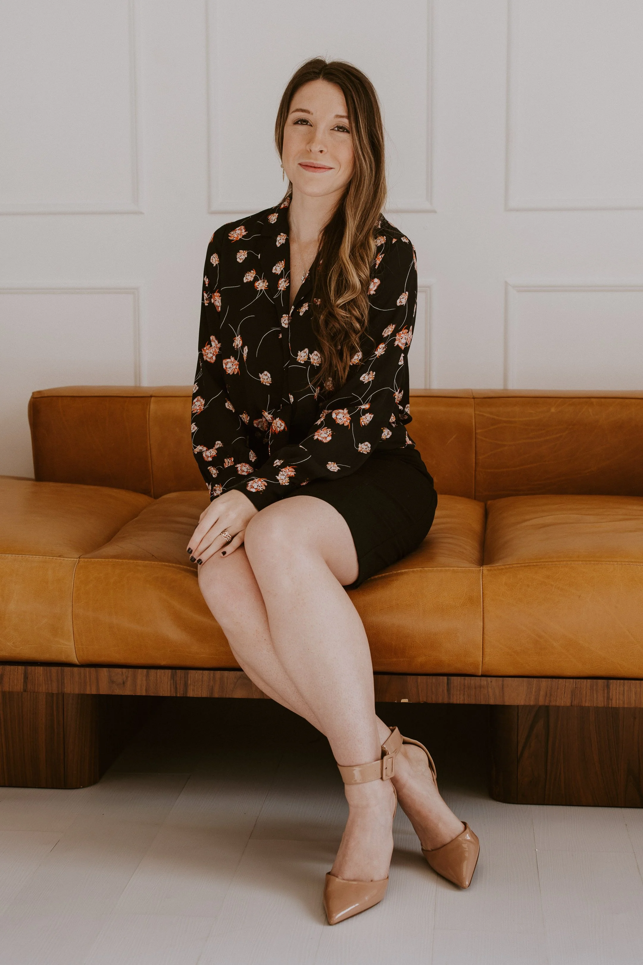 A woman with long brown hair sitting on a tan leather couch, wearing a black floral blouse, a black skirt, and beige high heels, in a room with white paneled walls.