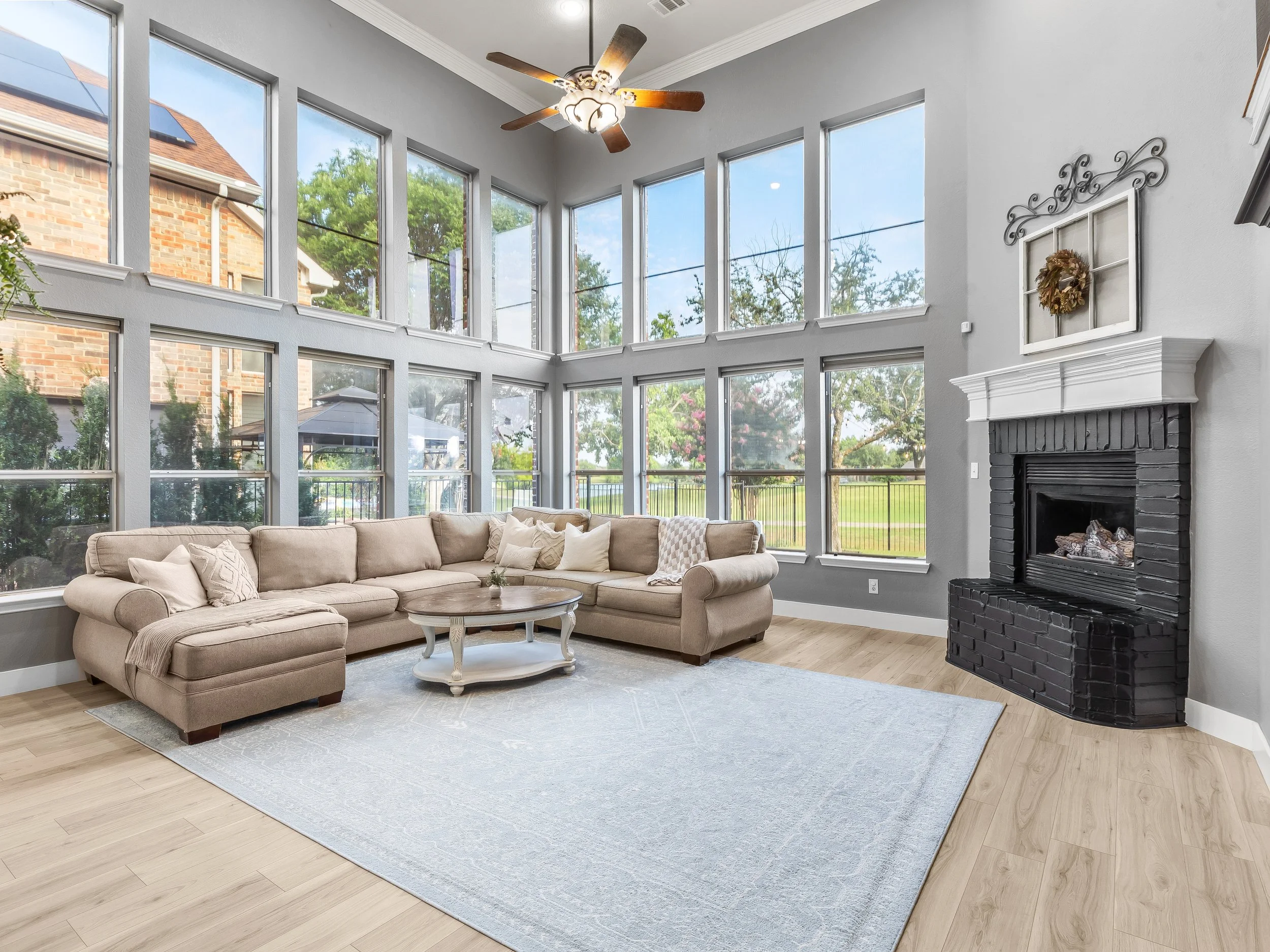 Living room with large windows, beige sectional sofa, light wood flooring, black brick fireplace, white popcorn ceiling fan, and a light-colored area rug.