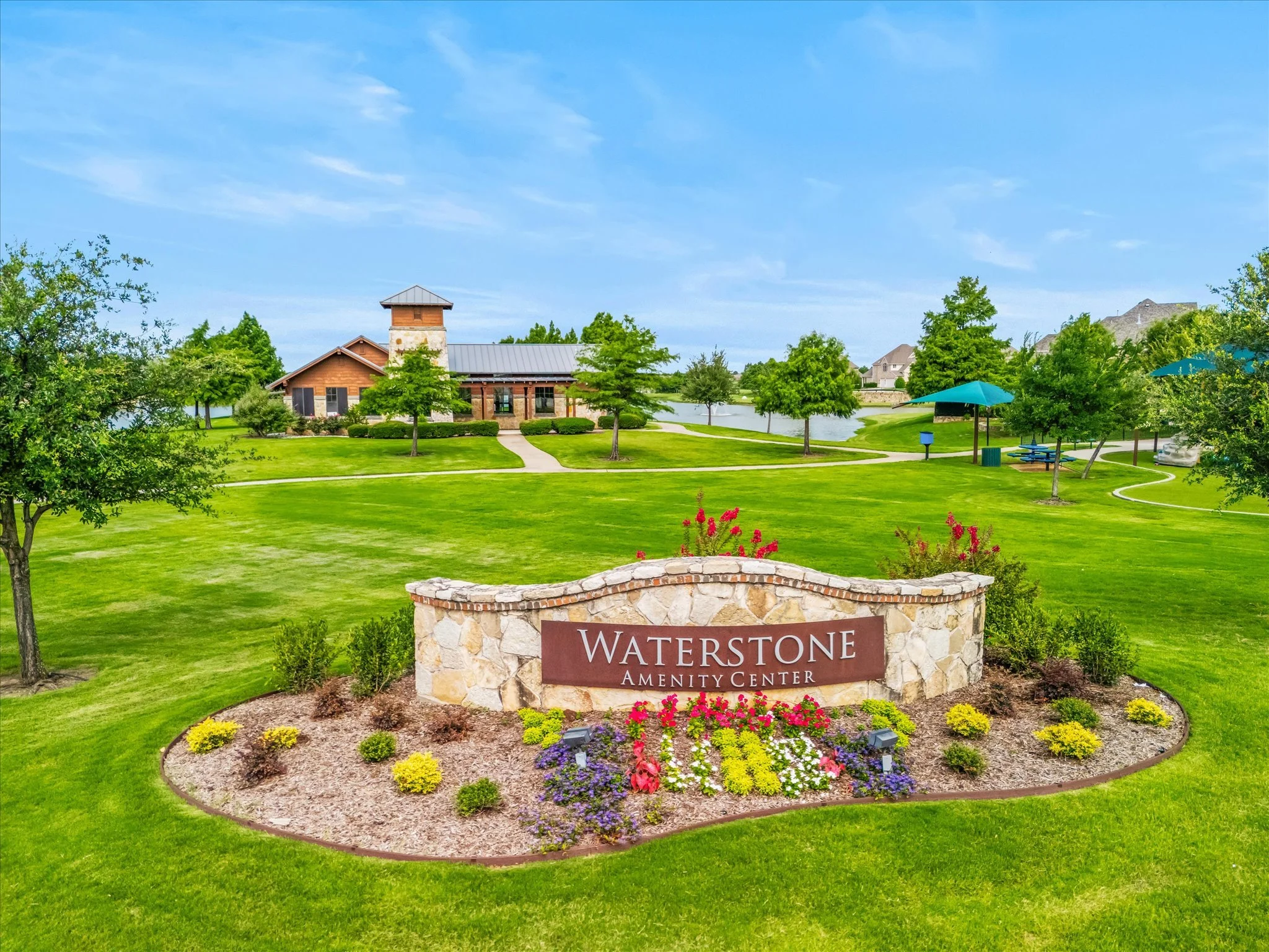 A landscaped park with a stone sign reading 'Waterstone Amenity Center' in front of a green lawn, trees, and a building with a tall tower, under a blue sky.