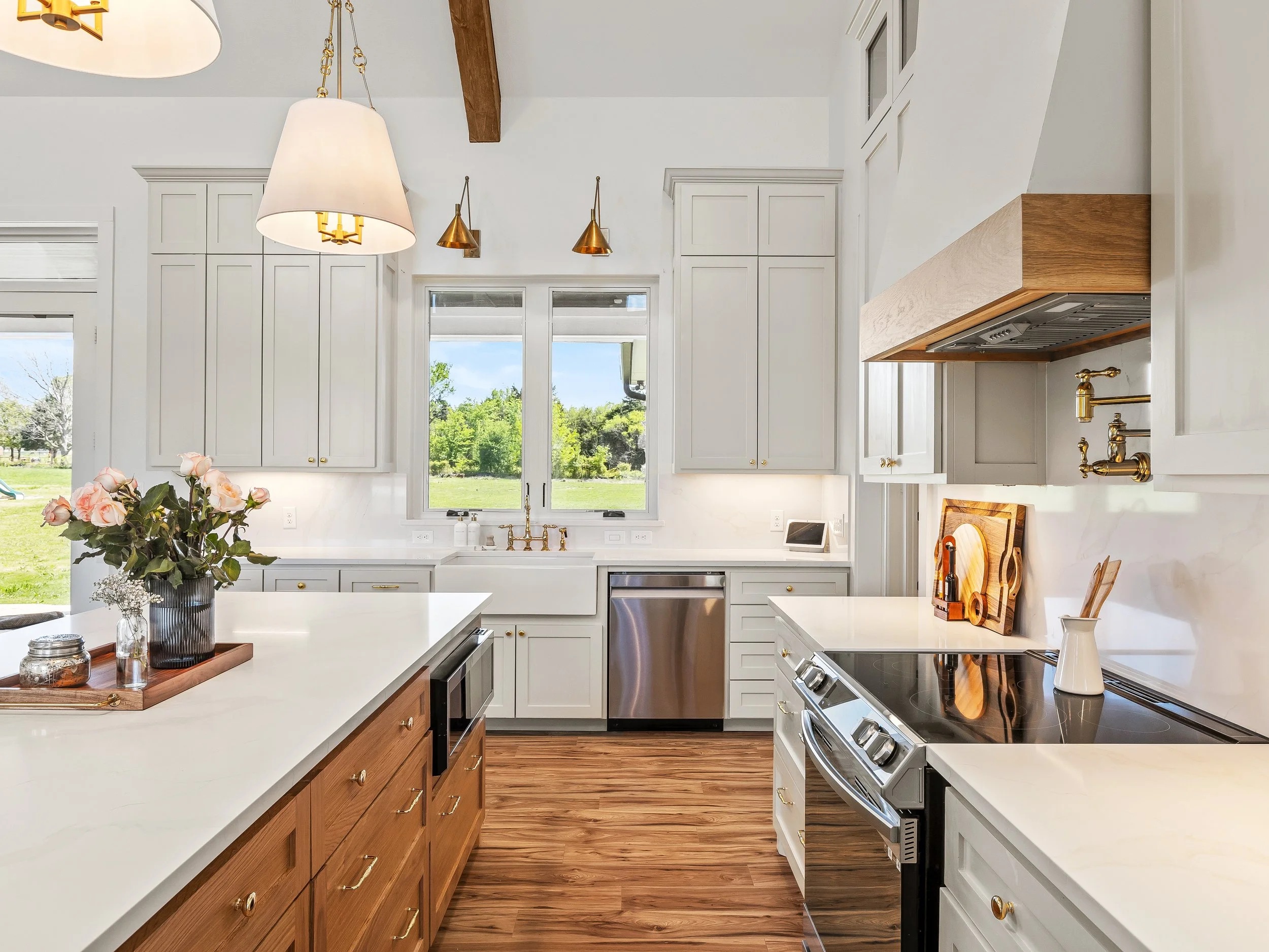 Modern kitchen with white cabinets, wooden countertops, stainless steel dishwasher, black stove, wooden island, hanging light fixtures, and a window with green trees outside.