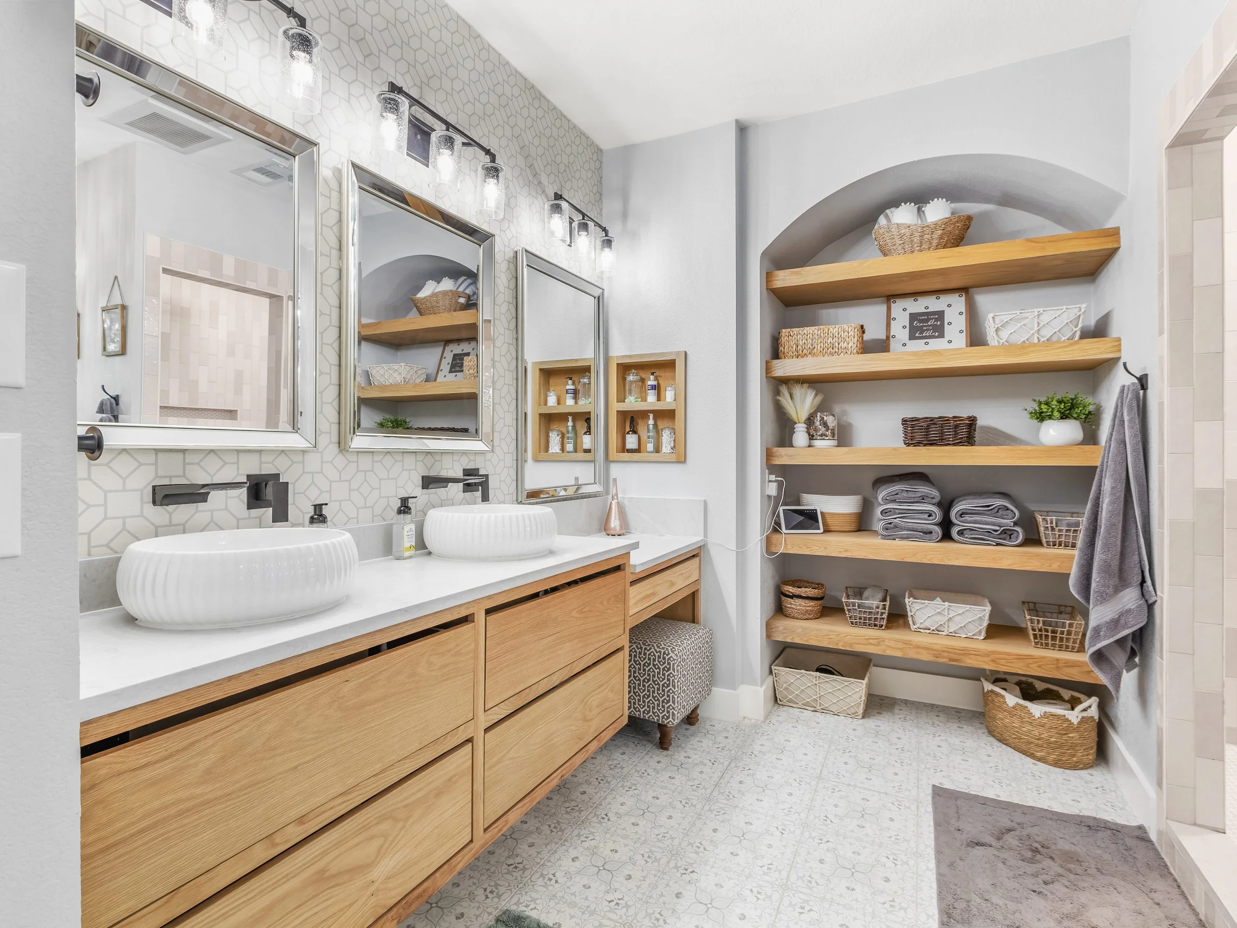 Bathroom with two vessel sinks, mirrored medicine cabinets, hexagon tile backsplash, open wooden shelves with baskets and towels, and a small stool.