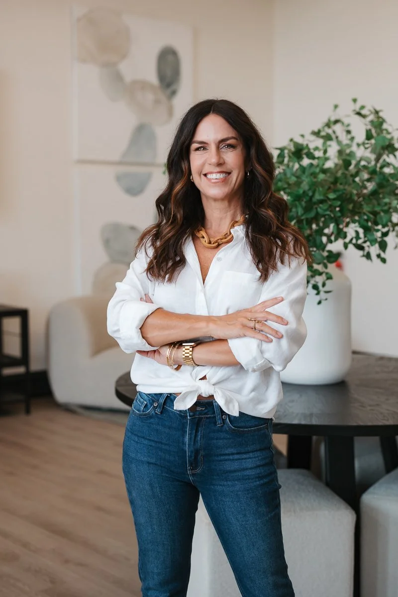 A woman with wavy brown hair, wearing a white knotted blouse and blue jeans, smiling and standing with arms crossed in a modern, well-lit living room with a large green plant and abstract artwork in the background.