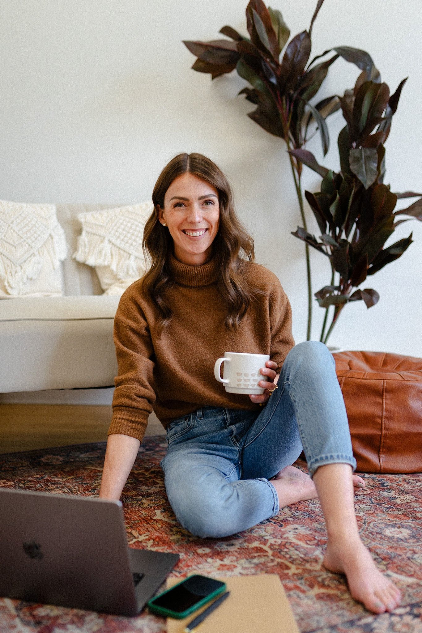 Michelle Ham sitting on a rug holding a coffee cup, with a laptop, smartphone, and notebook nearby, in a cozy room with plants and a white sofa. Michelle Ham offers virtual therapy sessions.