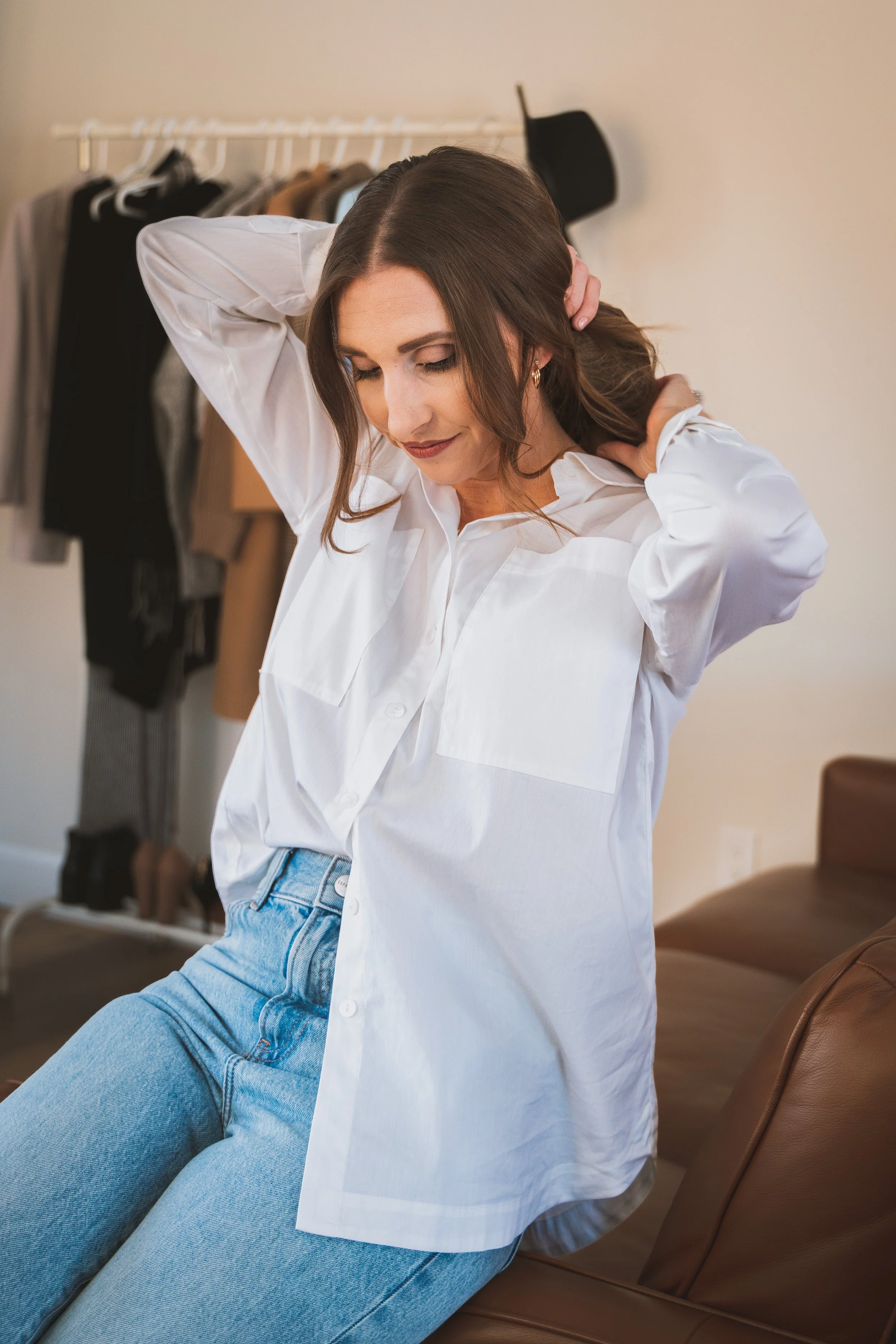 A woman with brown hair, wearing a white blouse and light blue jeans, sitting on a brown leather chair with her hands behind her head, in a room with a clothing rack in the background.