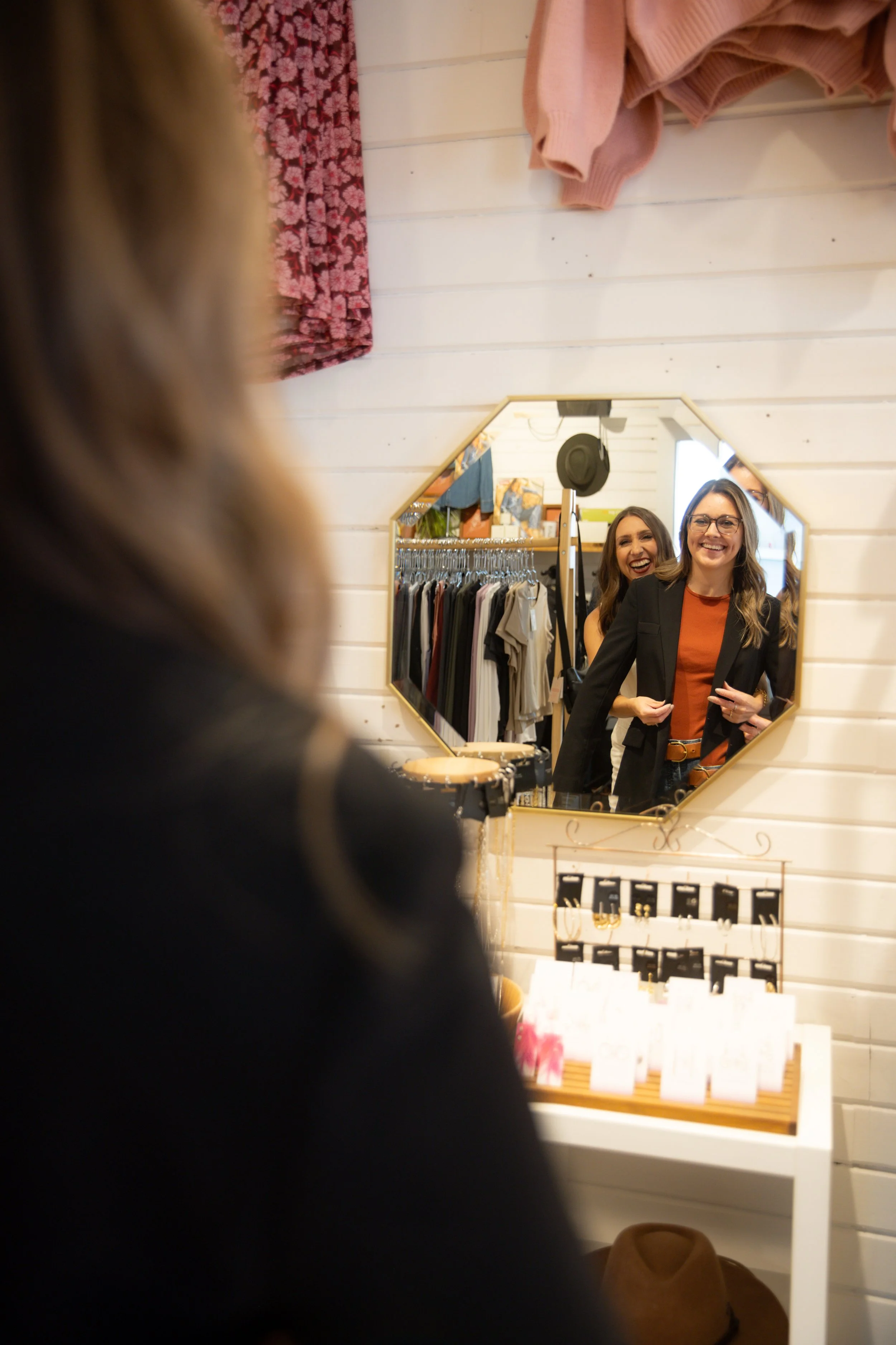Two women shopping in a clothing store, looking at their reflection in a wall-mounted mirror. They are smiling and appear happy. Clothing items are visible in the background.