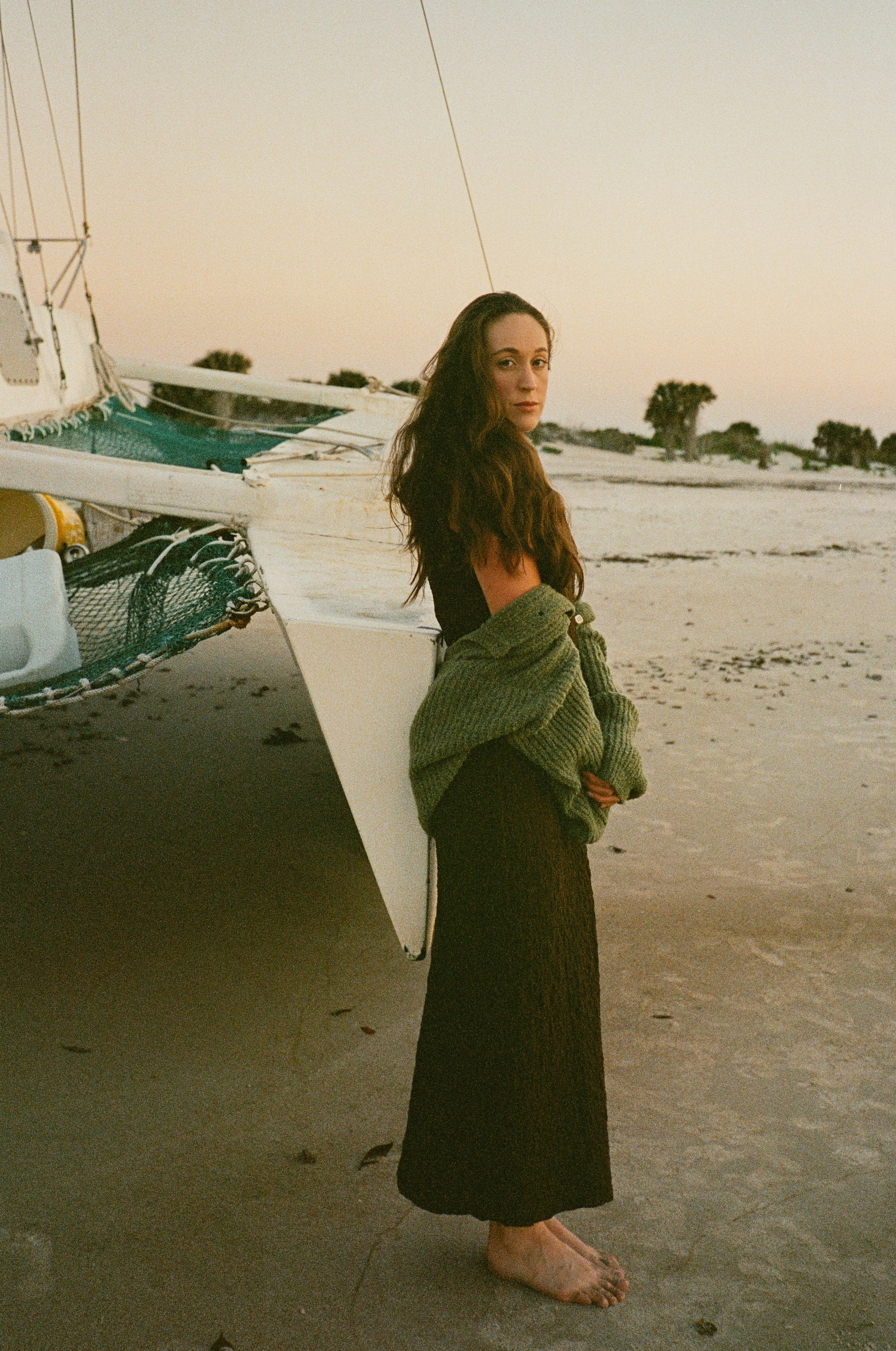 A woman with long brown hair standing barefoot on a beach next to a small boat at sunset, wearing a black dress and a green sweater tied around her waist.
